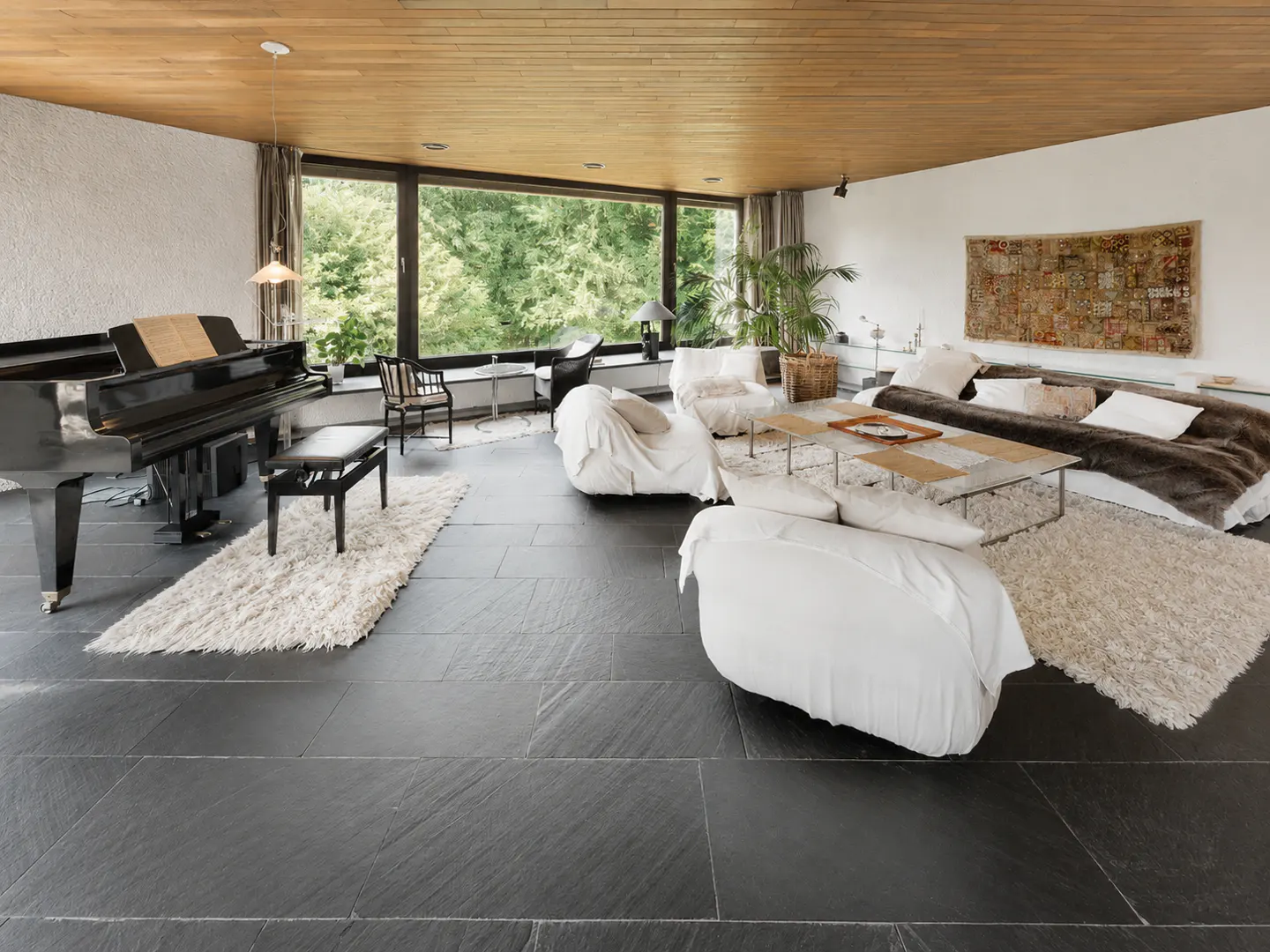 Modern living room with black slate floors, a grand piano, white lounge furniture, and large windows overlooking greenery.