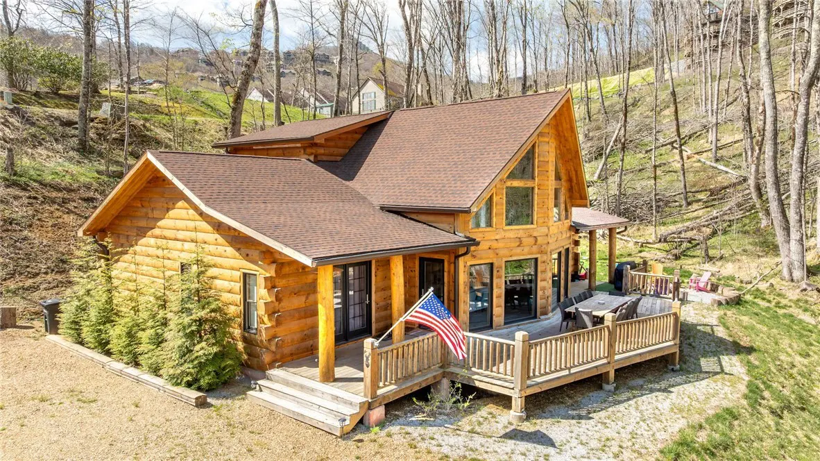 A two-story log cabin with a brown roof and a wooden deck featuring an American flag, set in a wooded mountain landscape.