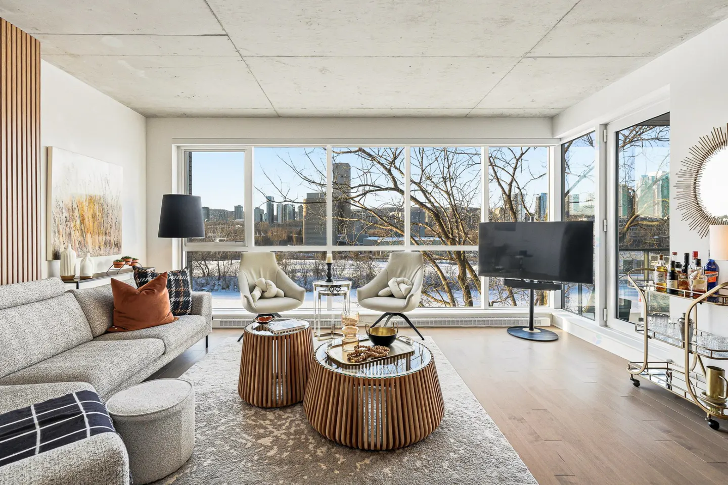 Bright living room with a gray sofa, two chairs, and a large window showing a city view. A bar cart stands near the window.