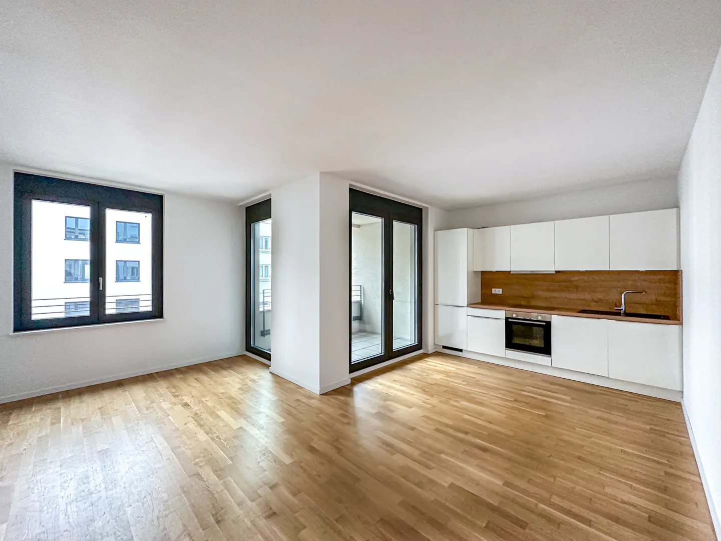 Bright, modern apartment with wood floors, white walls, and a kitchen with white cabinets. Large windows offer city views.