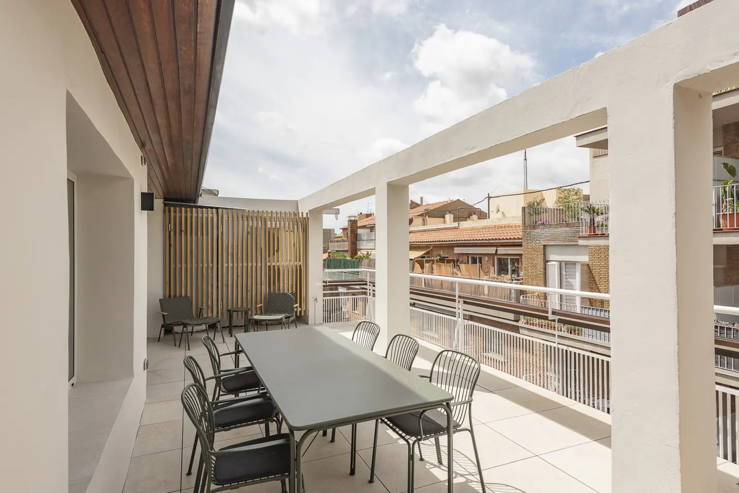 A rooftop patio with a long gray table and chairs, a bamboo screen, and a view of nearby buildings.