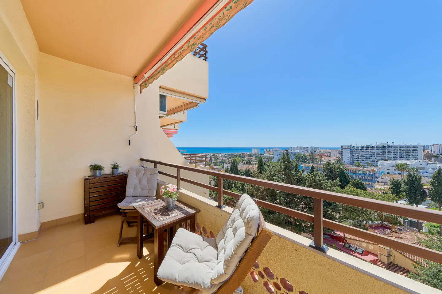 Balcony view with furniture overlooking a city and the ocean. Two chairs, a table, and a wooden cabinet are on the balcony.