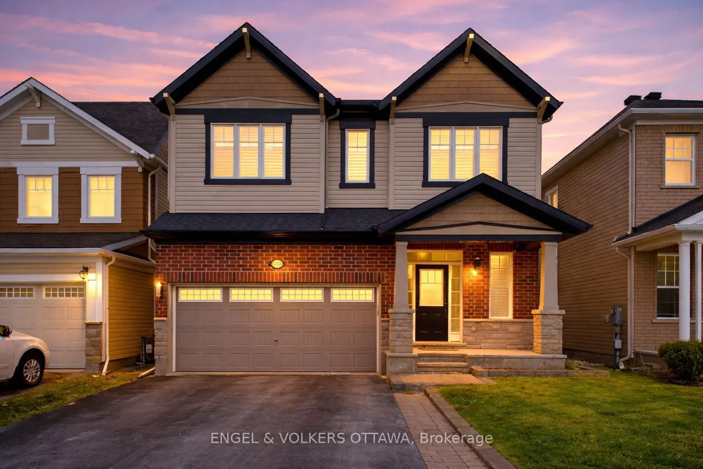 Two-story house with brick and beige siding, black roof, and a two-car garage at dusk. Windows are lit.