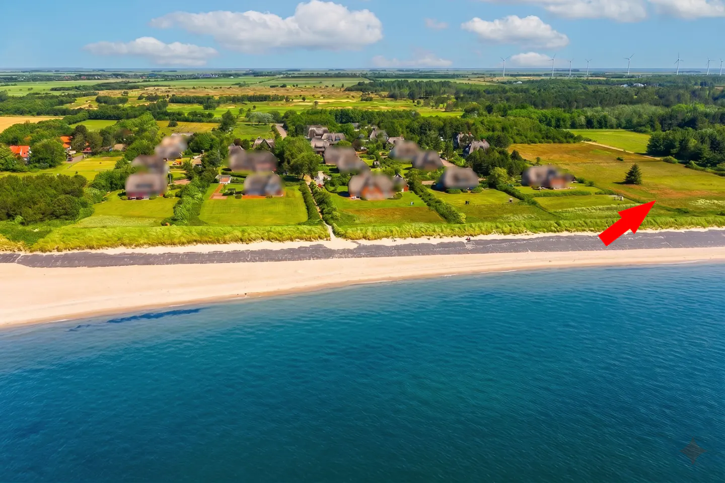 Aerial view of beach houses on a sandy shore with blue water. Green fields and wind turbines are in the background.