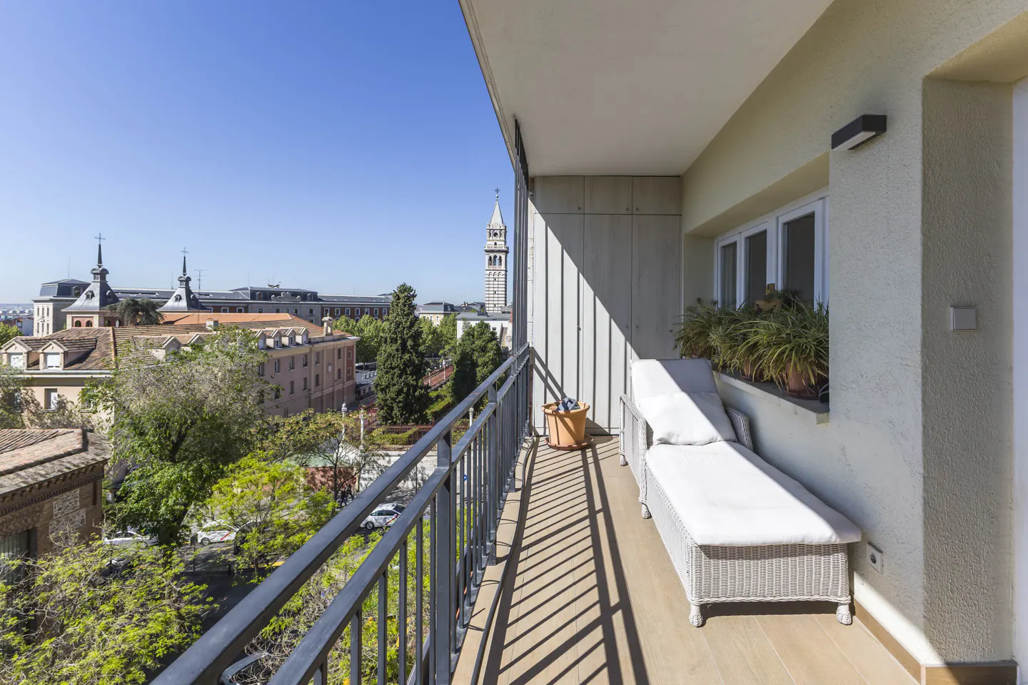 Balcony view with a white wicker chaise lounge, potted plants, and a cityscape with a church tower in the background.