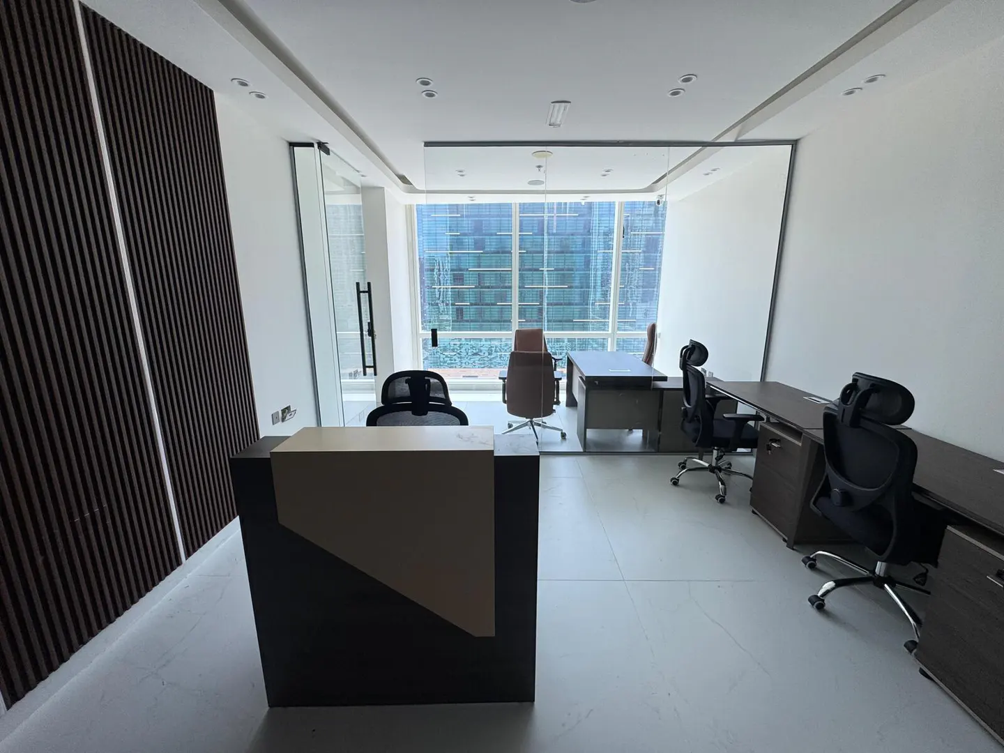 A modern office space with desks, chairs, and a reception area. A large window offers a city view.