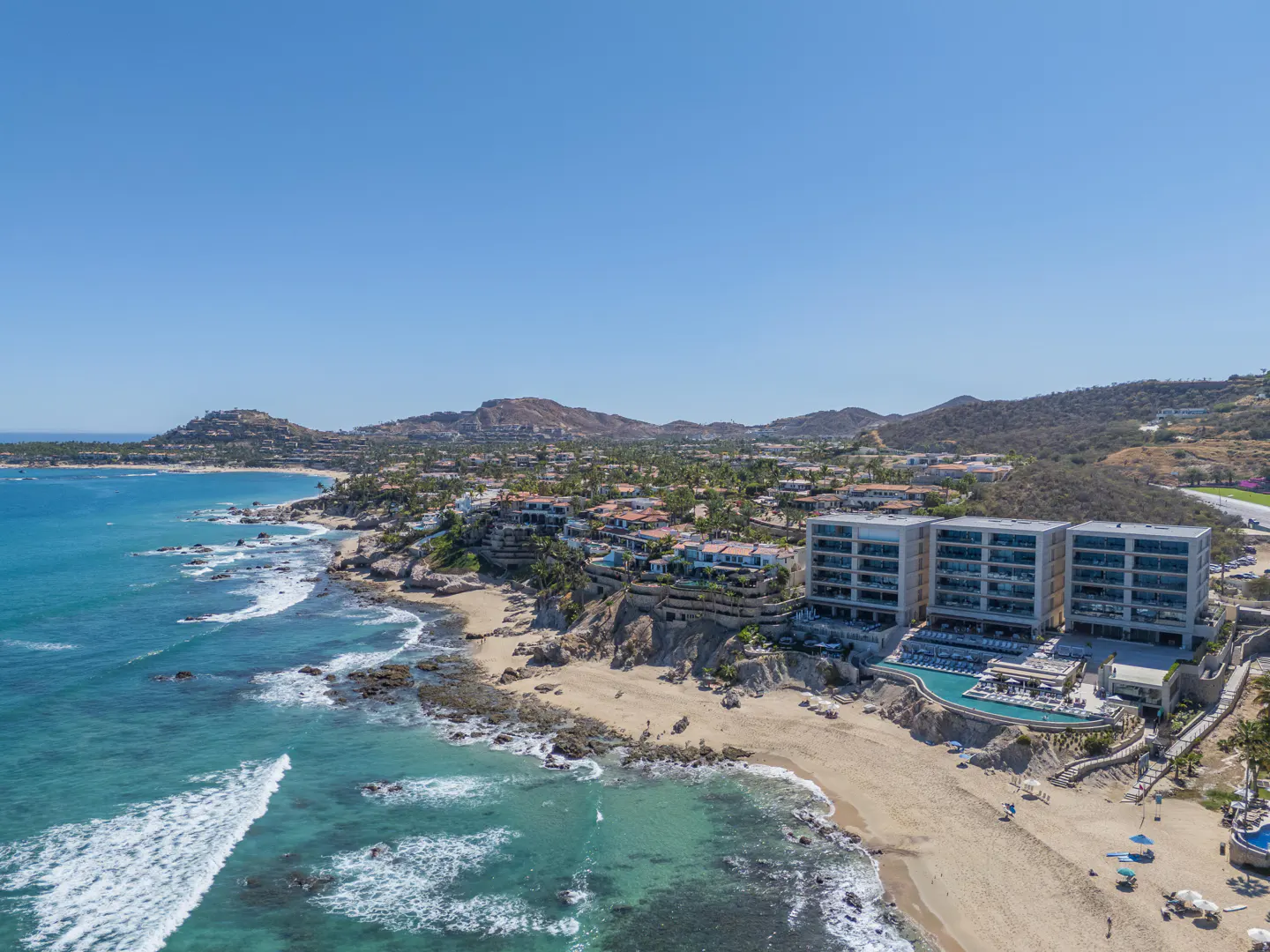Aerial view of a beachfront resort with turquoise ocean, sandy beach, and mountains in the background under a clear blue sky.