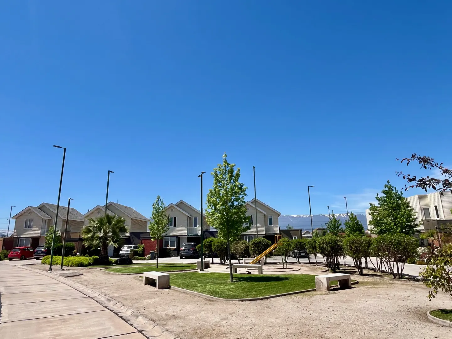 A suburban neighborhood with beige houses, green trees, a playground, and a clear blue sky.