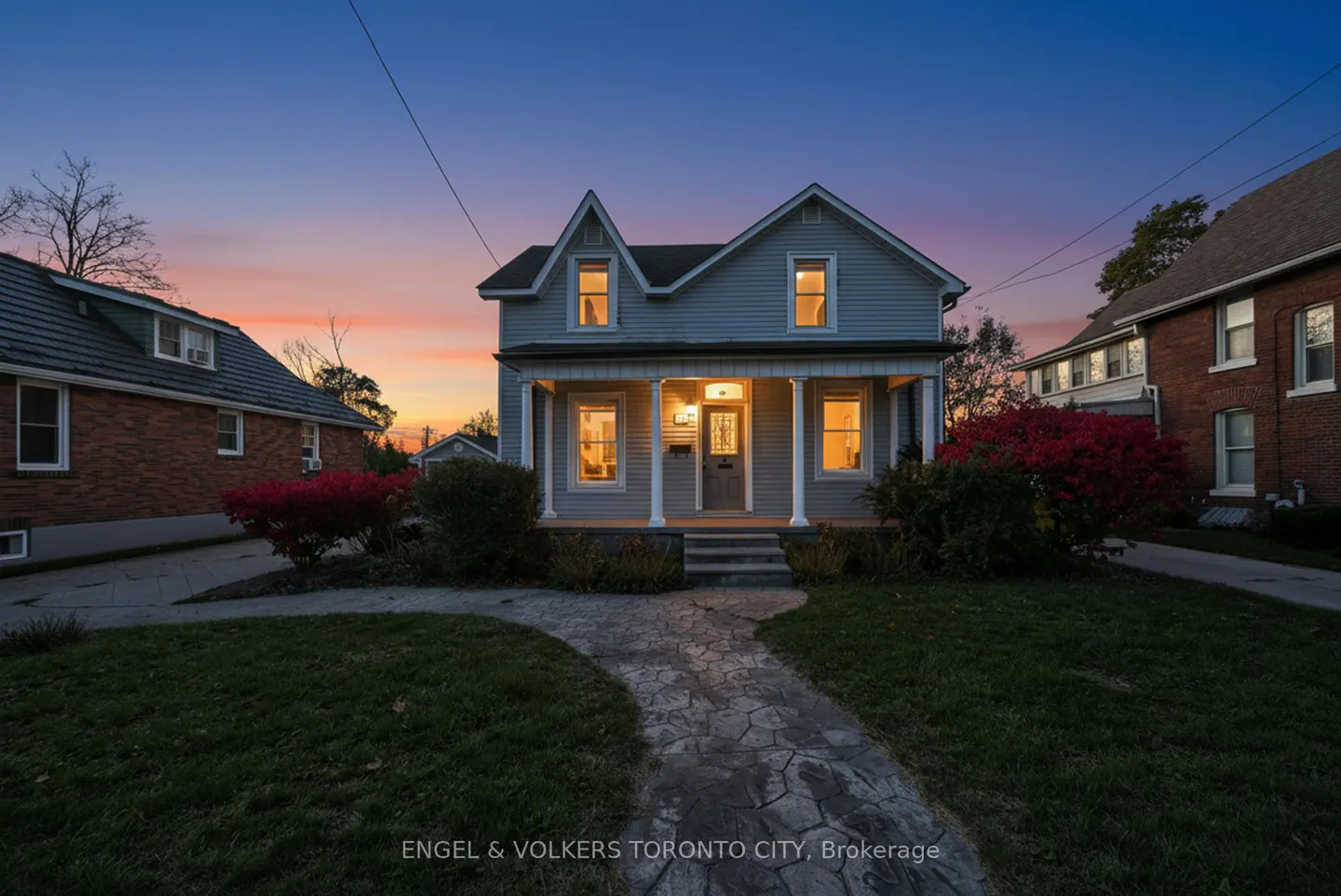 A two-story grey house with a front porch and glowing interior lights at sunset, featuring a stone walkway and lawn.