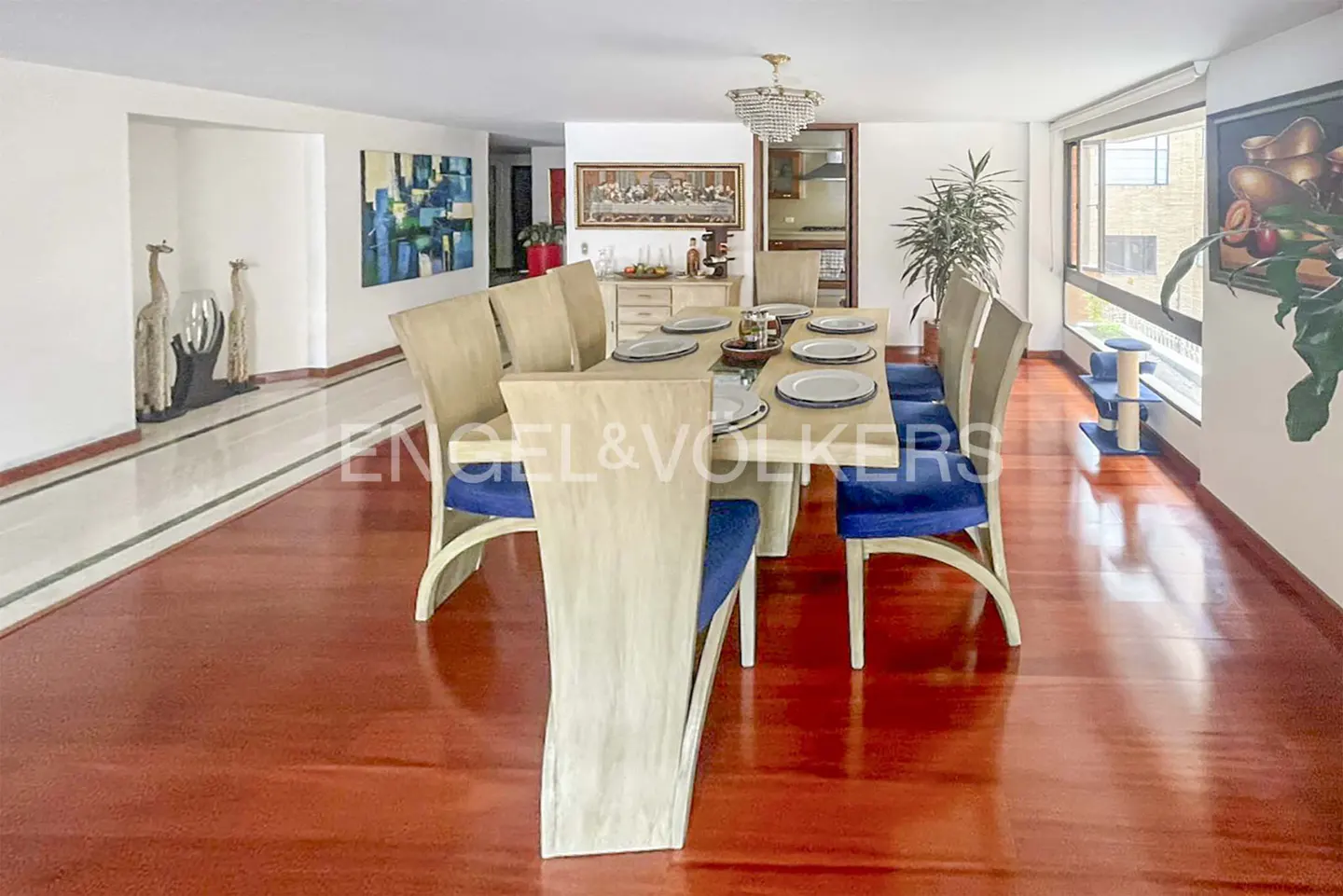 A dining room with a long table set for eight, light wood chairs with blue cushions, and a chandelier above.
