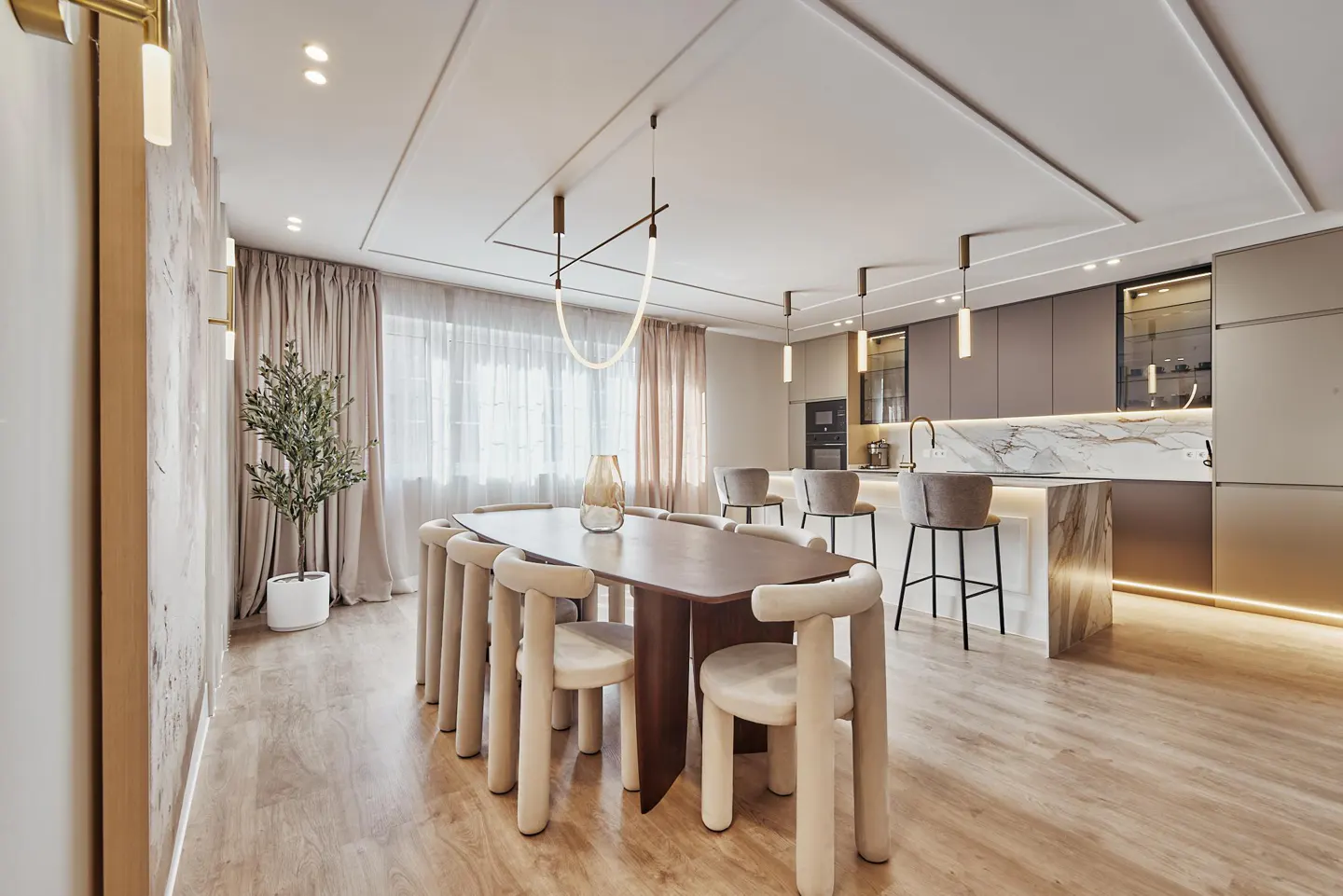 A modern, open-concept kitchen and dining area with light wood floors. A long, dark wood table is surrounded by beige chairs. A kitchen island with marble countertops is visible.