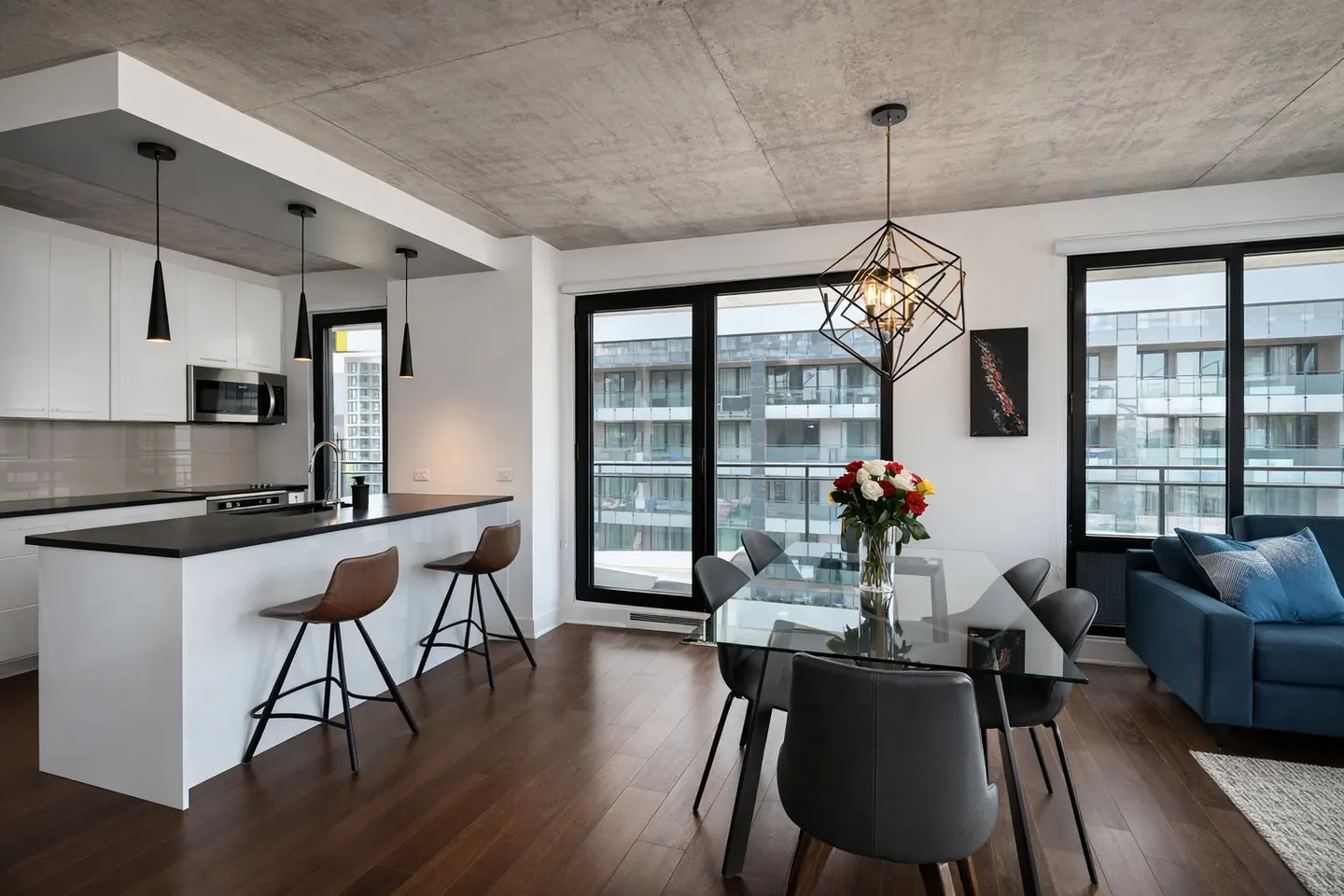 Bright, modern condo interior with a kitchen island, dining table with flowers, and a blue sofa near large windows.