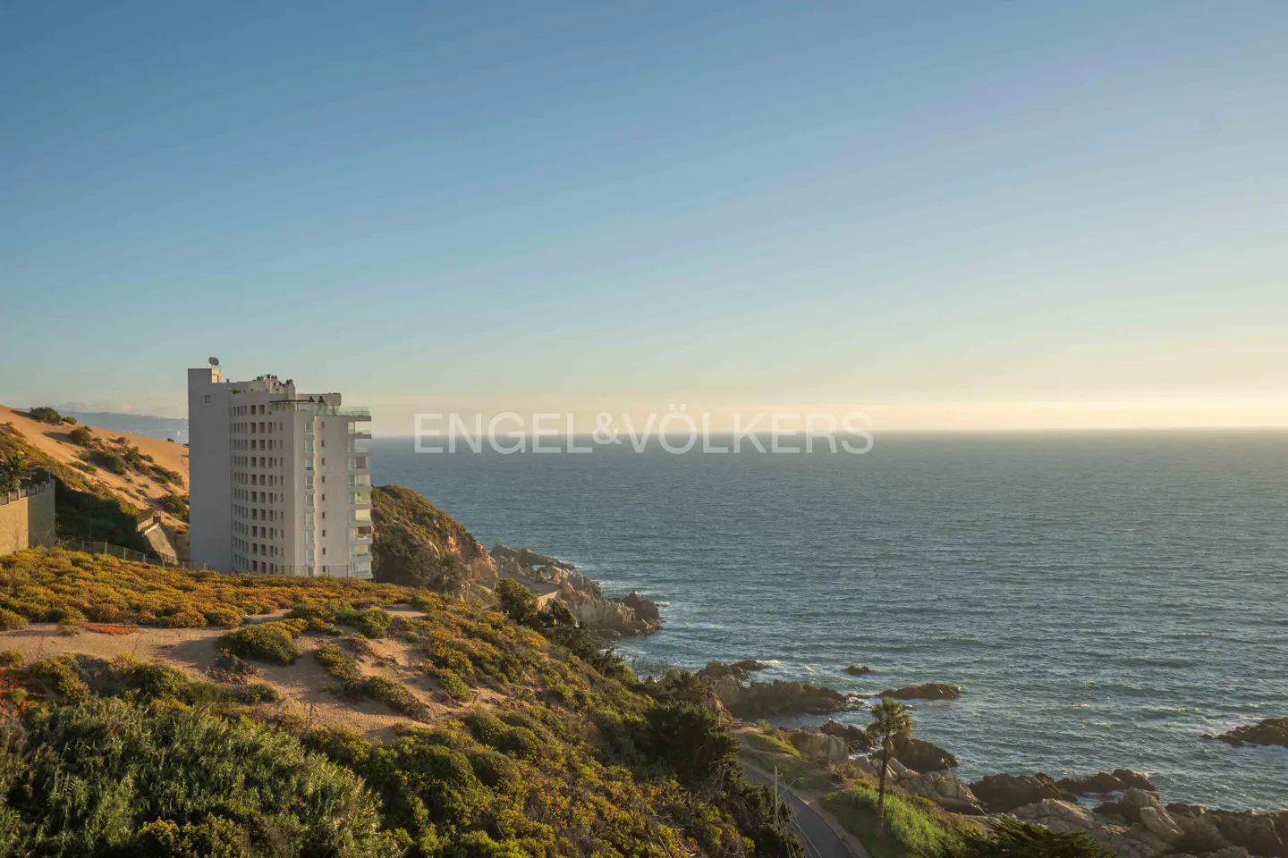 Ocean view with a tall white building on a green hillside under a blue sky. Engel & Völkers logo in the center.