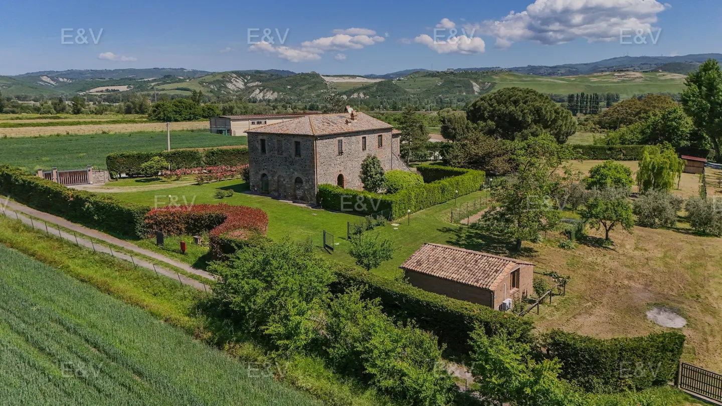Aerial view of a stone house with a red tile roof, surrounded by green lawns, trees, and a hedge fence in a rural setting.