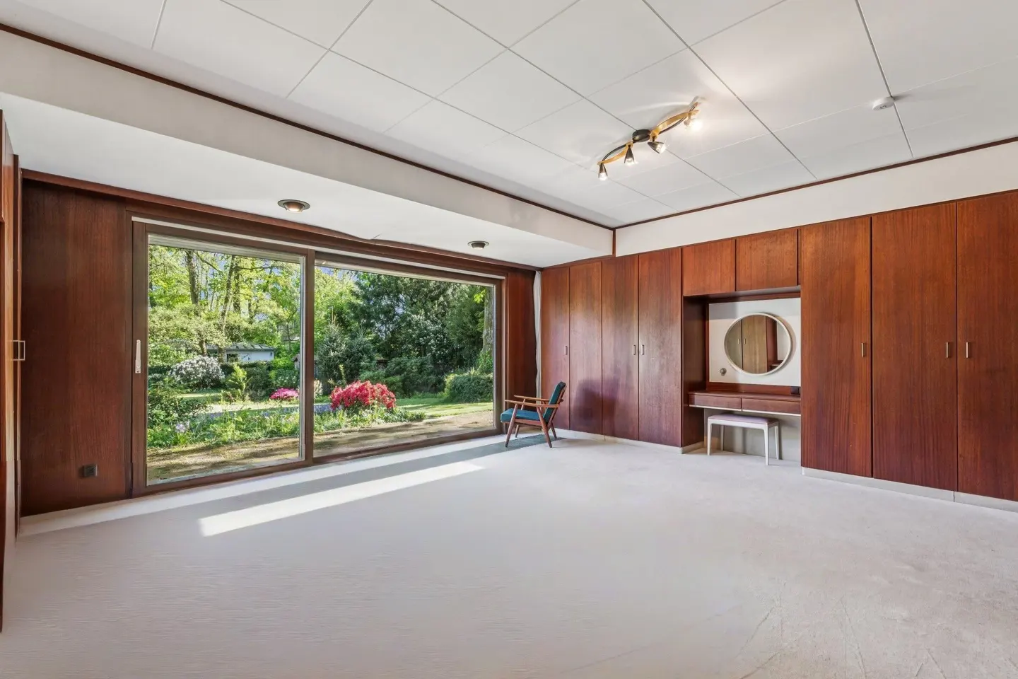 Bright bedroom with white carpet, wood paneled walls, and a large window overlooking a garden. A blue chair sits near the window.