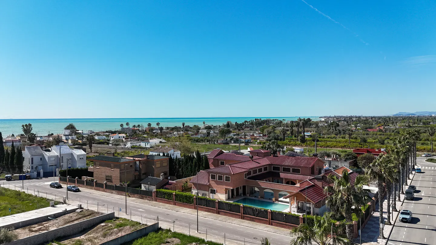 Aerial view of a large, red-roofed house with a pool, palm trees, and the ocean in the background under a clear blue sky.