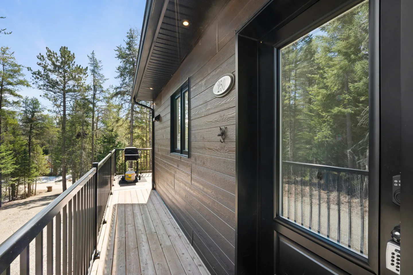 Exterior view of a modern brown house with a wooden deck, black railings, and a grill, surrounded by green trees.
