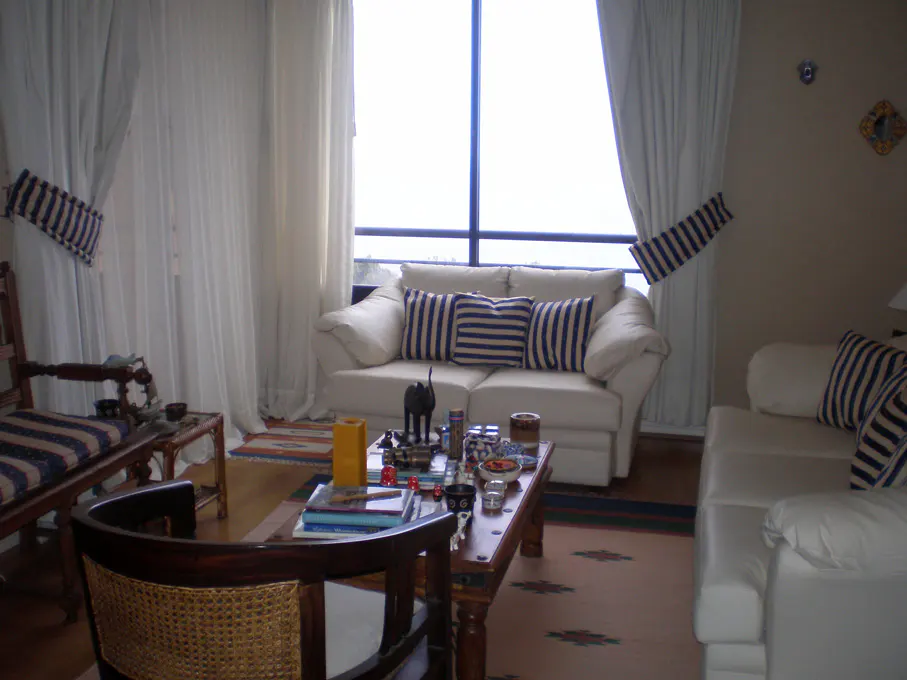 Living room with white sofas, striped pillows, and a wooden coffee table with books and decorative items. Large windows with white curtains.