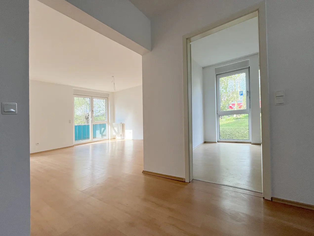 An interior view of a bright, empty apartment with light wood floors and white walls, seen through doorways.