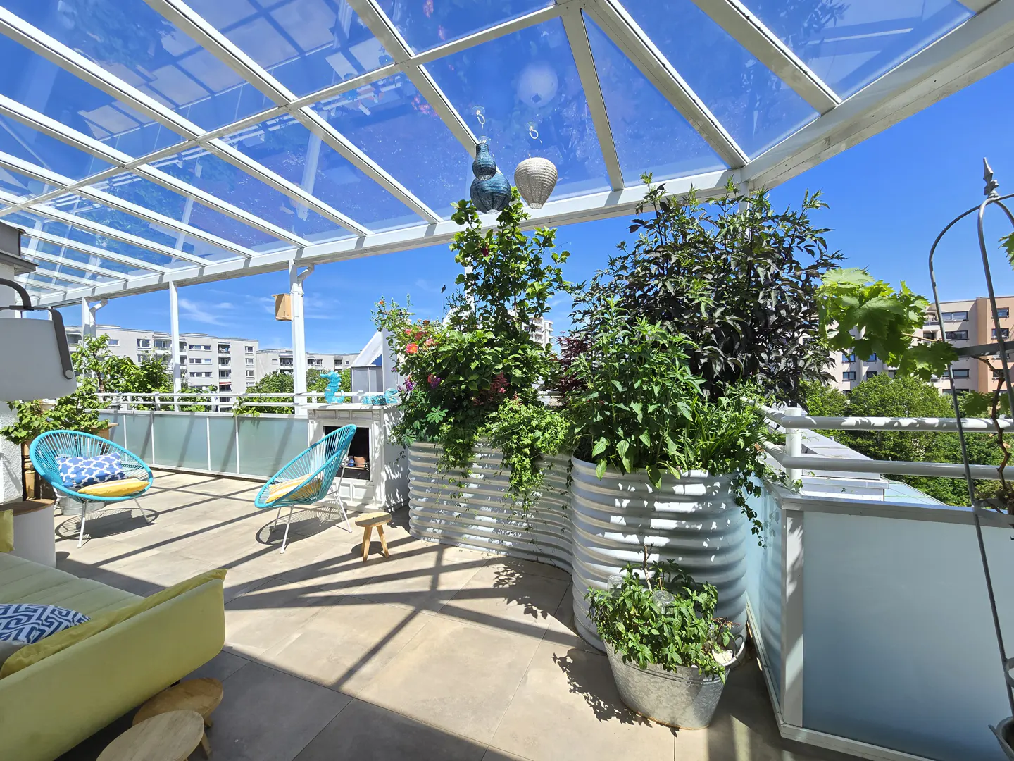 Sunny rooftop terrace with a glass roof, two blue chairs, lush potted plants, and a view of city buildings.