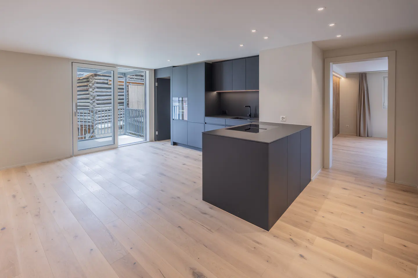 A modern kitchen with light wood floors, dark gray cabinets, and a kitchen island. Sliding glass doors lead to a balcony.