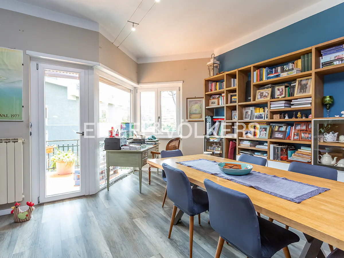 A bright dining room with a wooden table, blue chairs, and a large bookshelf against a blue wall. A desk sits near the open doors to a balcony.