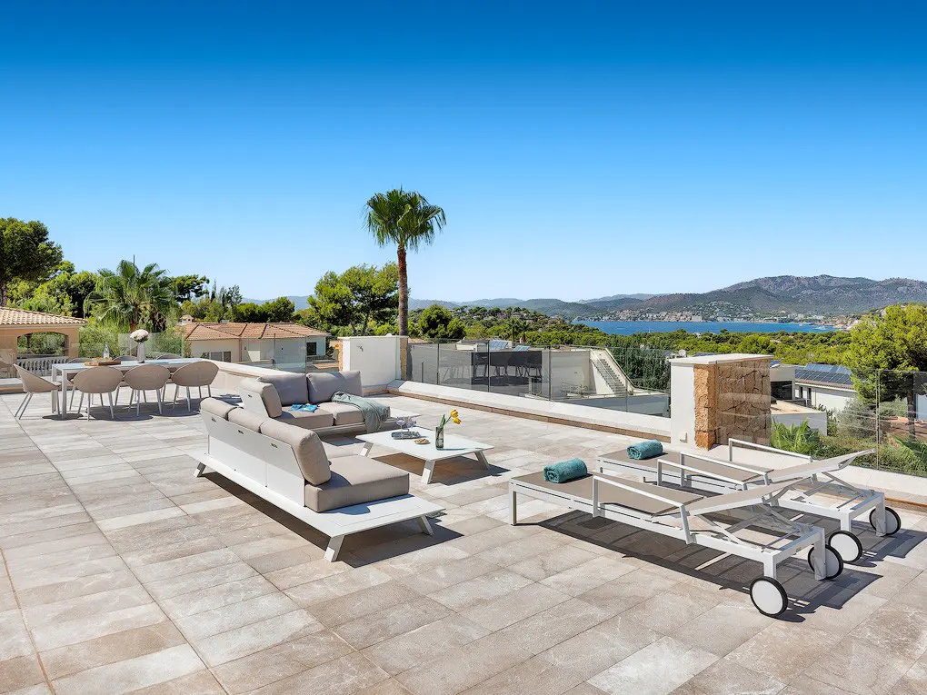 Luxury rooftop patio with lounge seating, dining table, and sun loungers overlooking a scenic view of the sea and mountains.