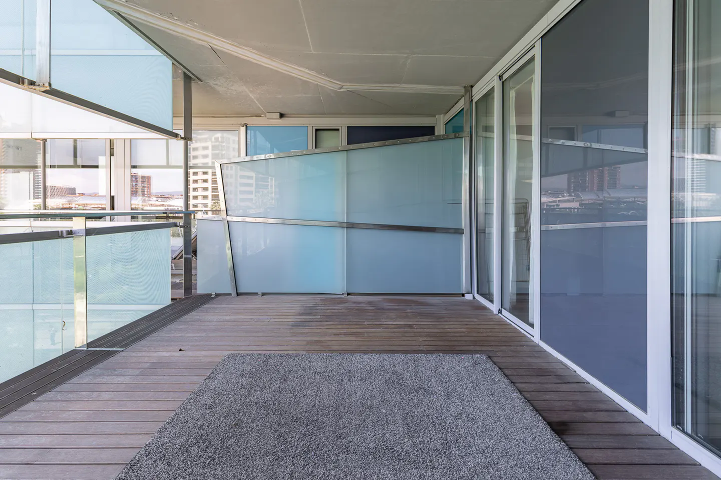Modern balcony with wood floor, gray rug, and blue glass panels. Sliding glass doors lead inside. City view in the background.
