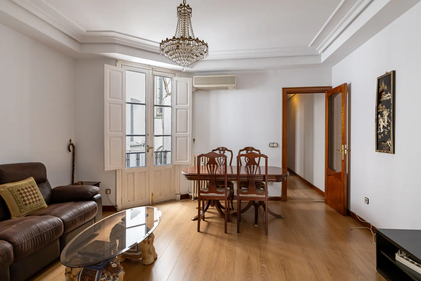 Living room with brown leather sofa, glass coffee table, dining table with chairs, and a crystal chandelier. White walls and wood floors.