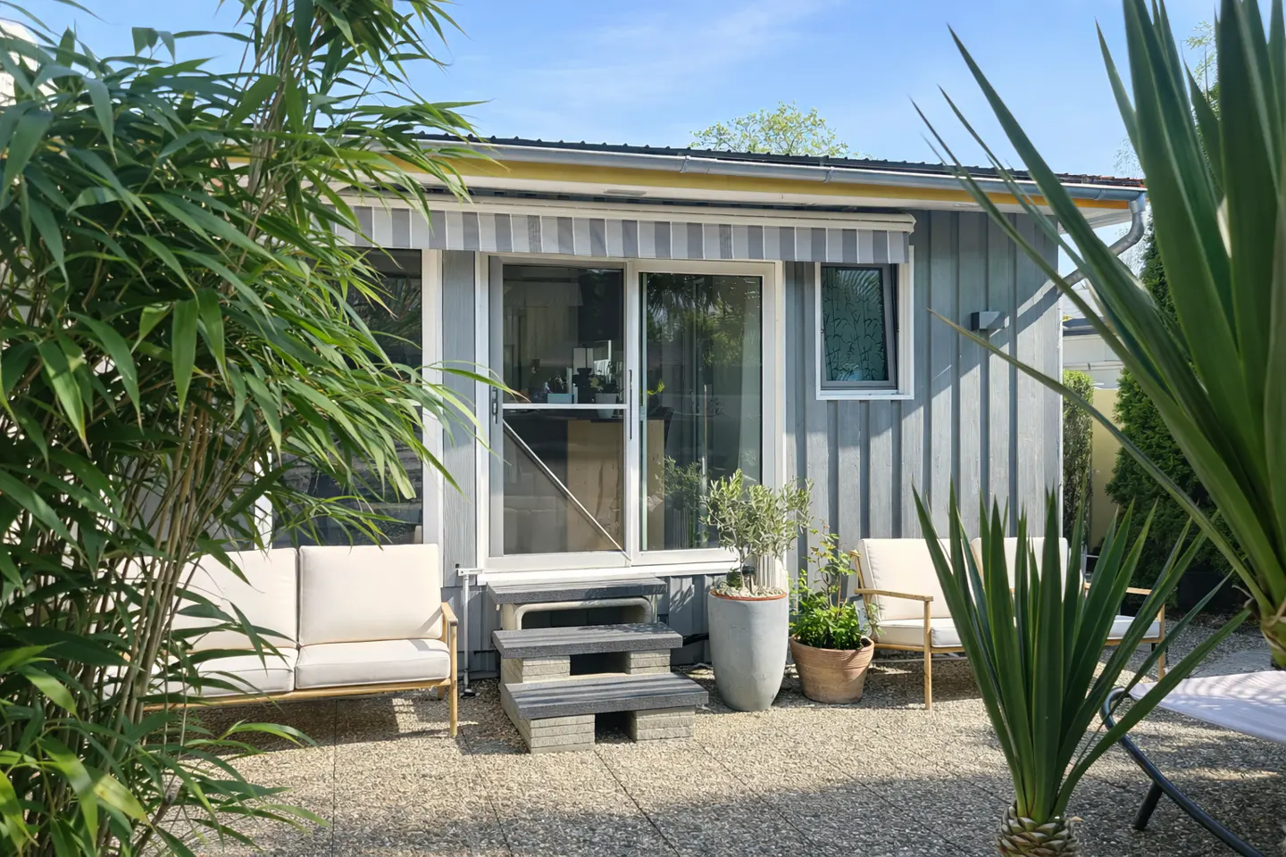 Patio with grey wood siding, sliding glass doors, striped awning, white outdoor furniture, and potted plants on gravel.
