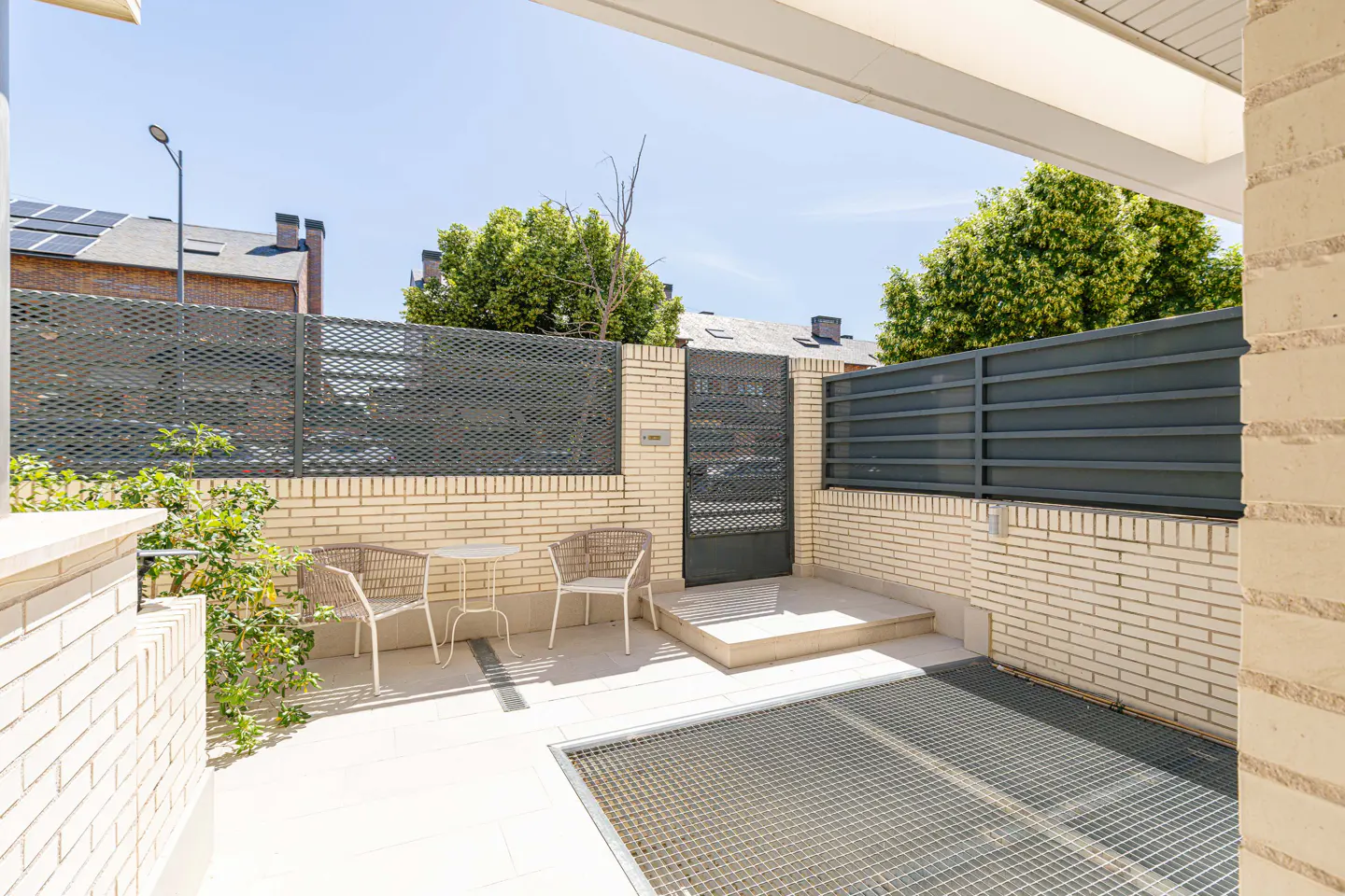 Outdoor patio with brick walls, metal fences, and a metal grate floor. A small table and two chairs sit on the patio.