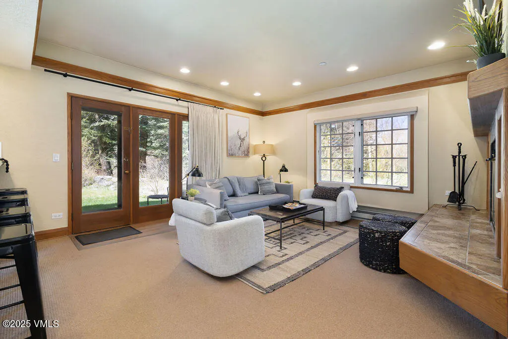 Living room with beige carpet, light yellow walls, and wood trim. A gray sofa and two white armchairs surround a coffee table on a patterned rug. French doors lead to a green lawn.