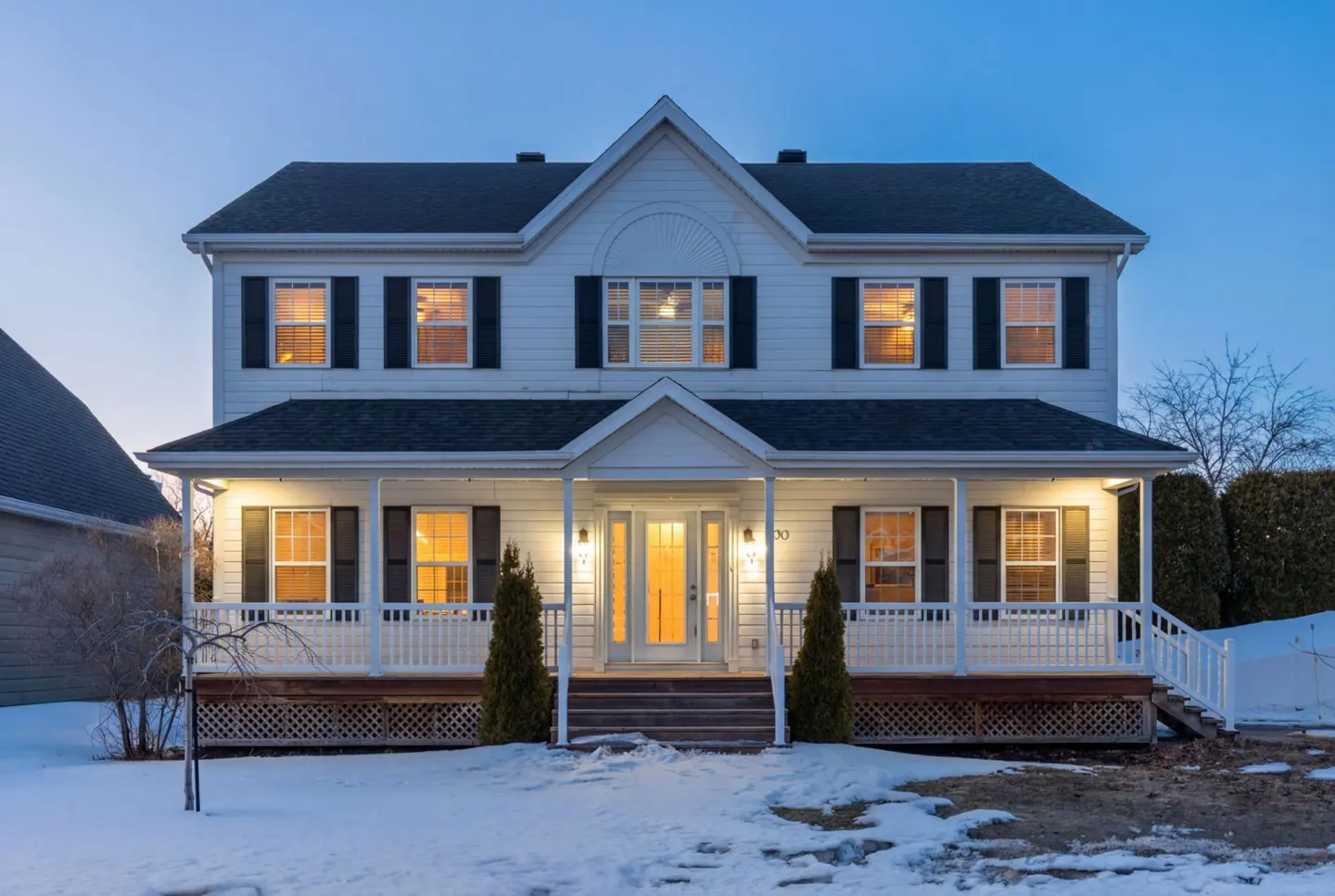 Two-story white house with black shutters and a porch, illuminated at dusk with snow on the ground.