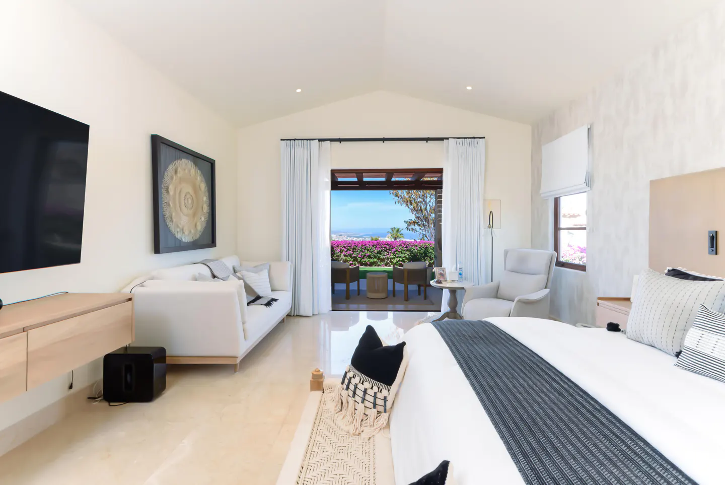 Bright bedroom with a white sofa, chair, and bed. A patio with pink flowers overlooks the ocean. A TV hangs on the wall.