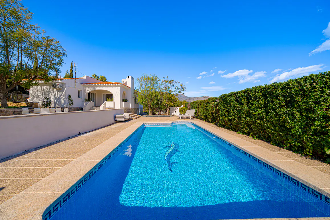 A bright blue swimming pool with a dolphin mosaic, surrounded by a stone patio, a white house, and green hedges under a clear blue sky.
