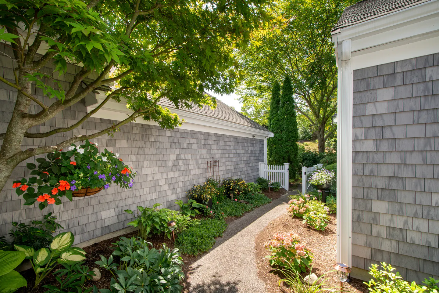 Landscaped walkway between two gray shingle houses leads to a white picket fence. Hanging flower basket on the left.
