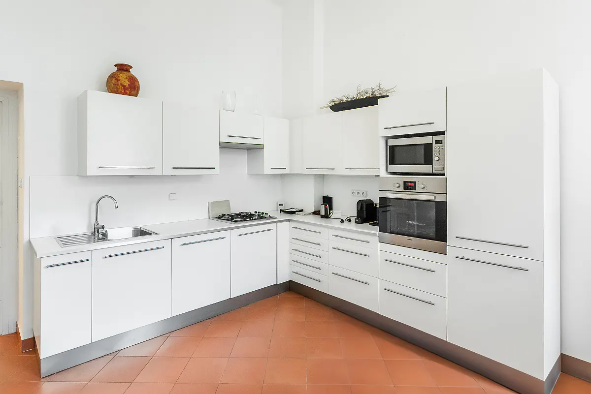 Bright kitchen with white cabinets, stainless steel appliances, and terracotta tile flooring. A decorative vase sits atop the cabinets.