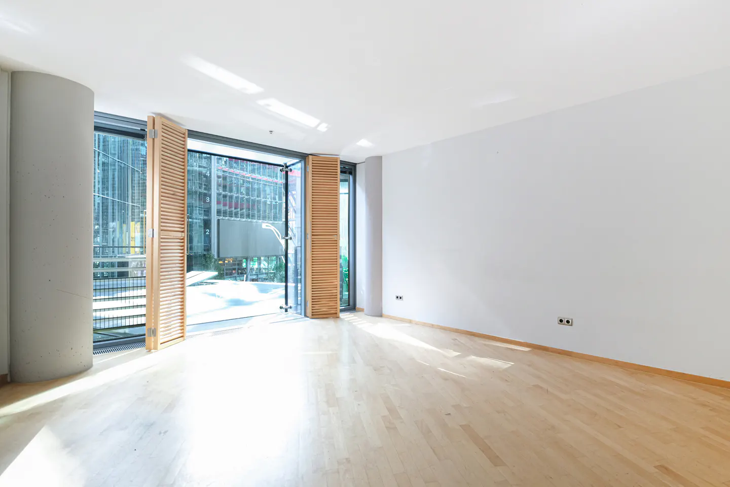 Bright, empty room with light wood floors, white walls, and open shuttered doors leading to a city view.