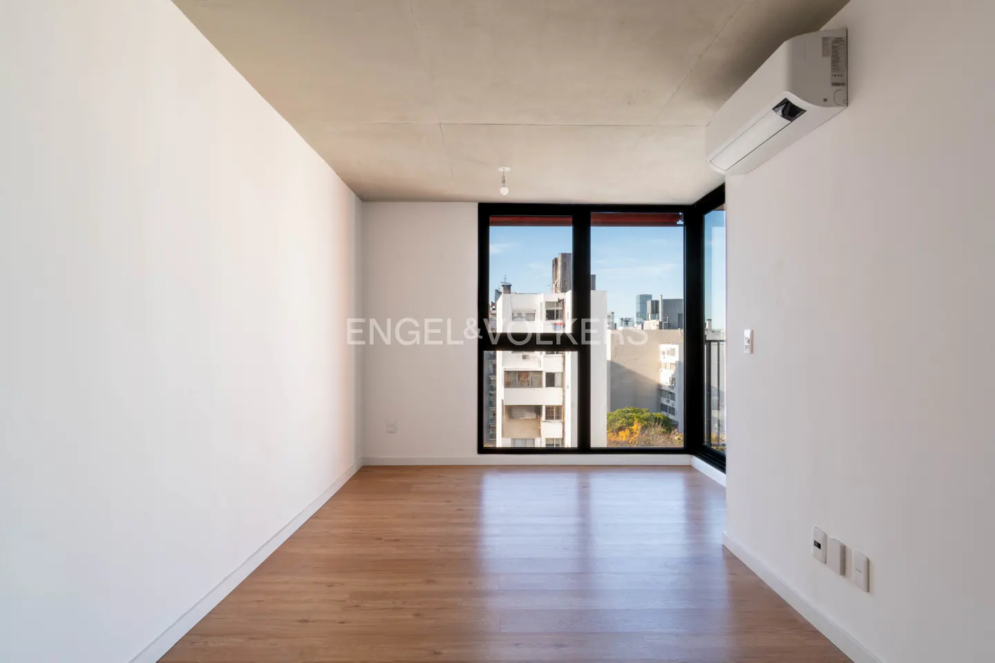 Bright, empty room with wood floors, white walls, and a black-framed window showing a city view. An air conditioner is mounted high on the wall.