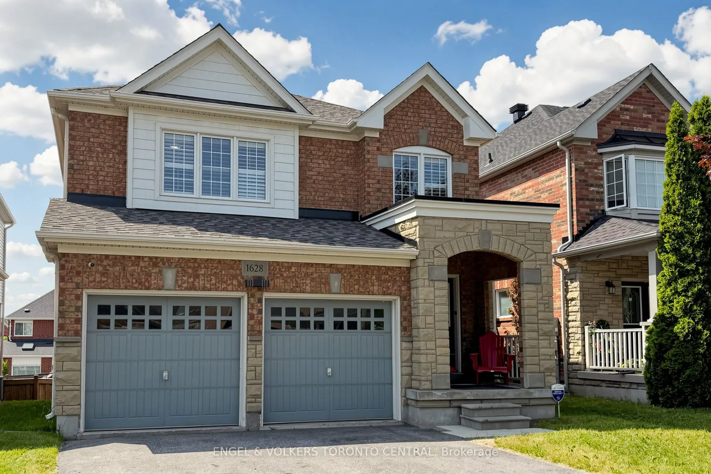 Two-story brick house with a double grey garage, stone porch, and a red chair under a blue sky with white clouds.