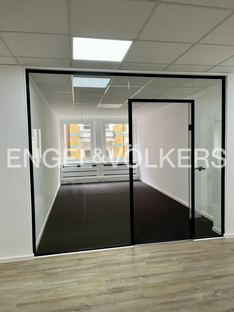Office space with glass-walled rooms, black trim, and light wood floors. The interior has dark carpet and white walls. Windows are visible in the background.