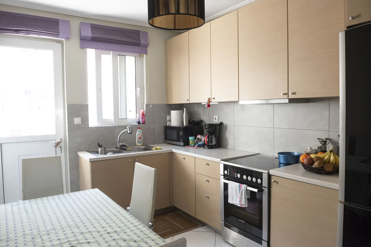 Bright kitchen with light wood cabinets, gray backsplash, and stainless steel appliances. A table with a checkered cloth is in the foreground.