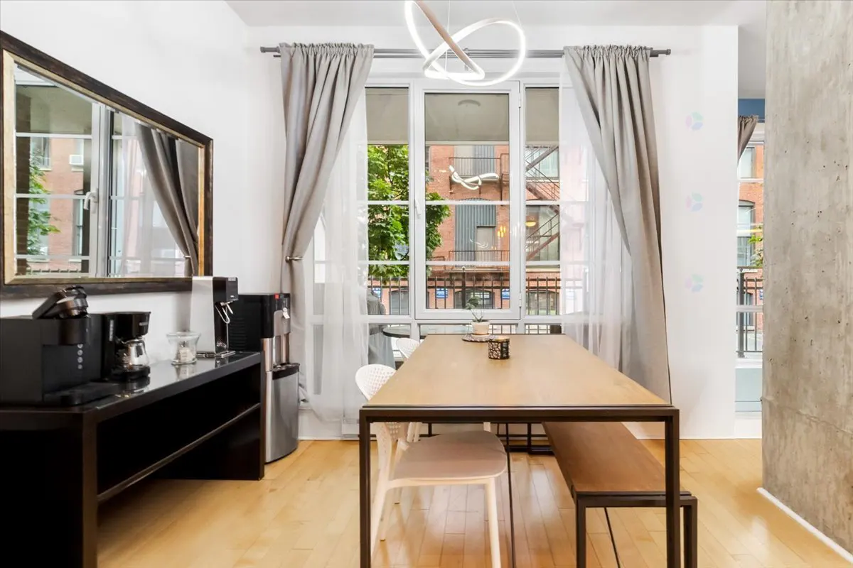 Bright dining area with a wood table, bench, and chairs. Large window with gray curtains and a view of a brick building. Black console with coffee makers.