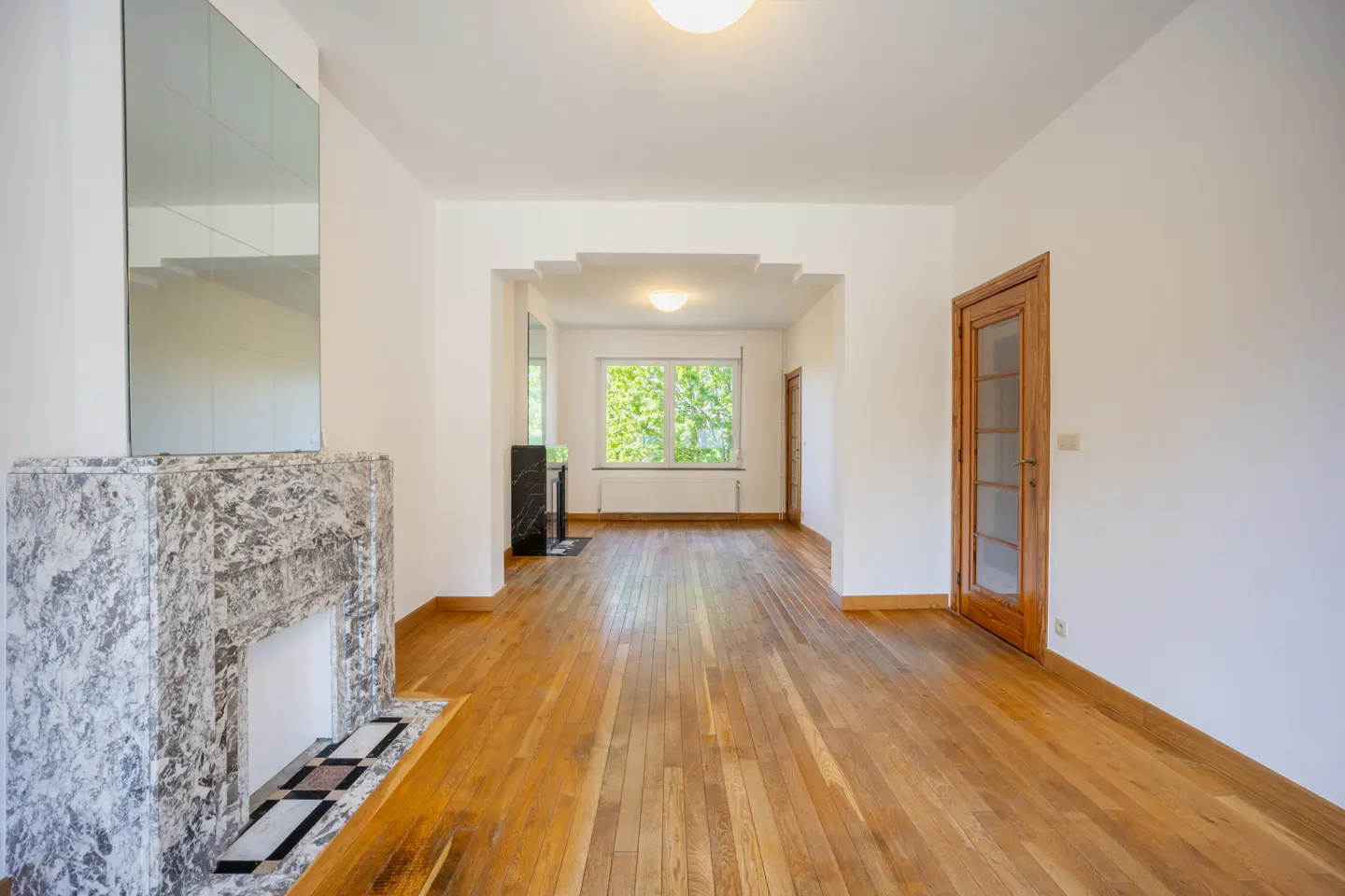 Bright, empty room with hardwood floors, white walls, and a marble fireplace. A doorway leads to another room with a window.