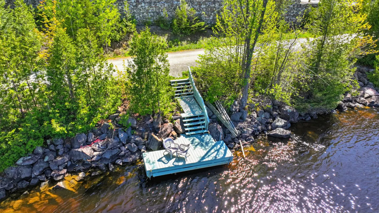 Aerial view of a wooden dock with two chairs, connected by stairs to a rocky shoreline with lush green trees and a path.