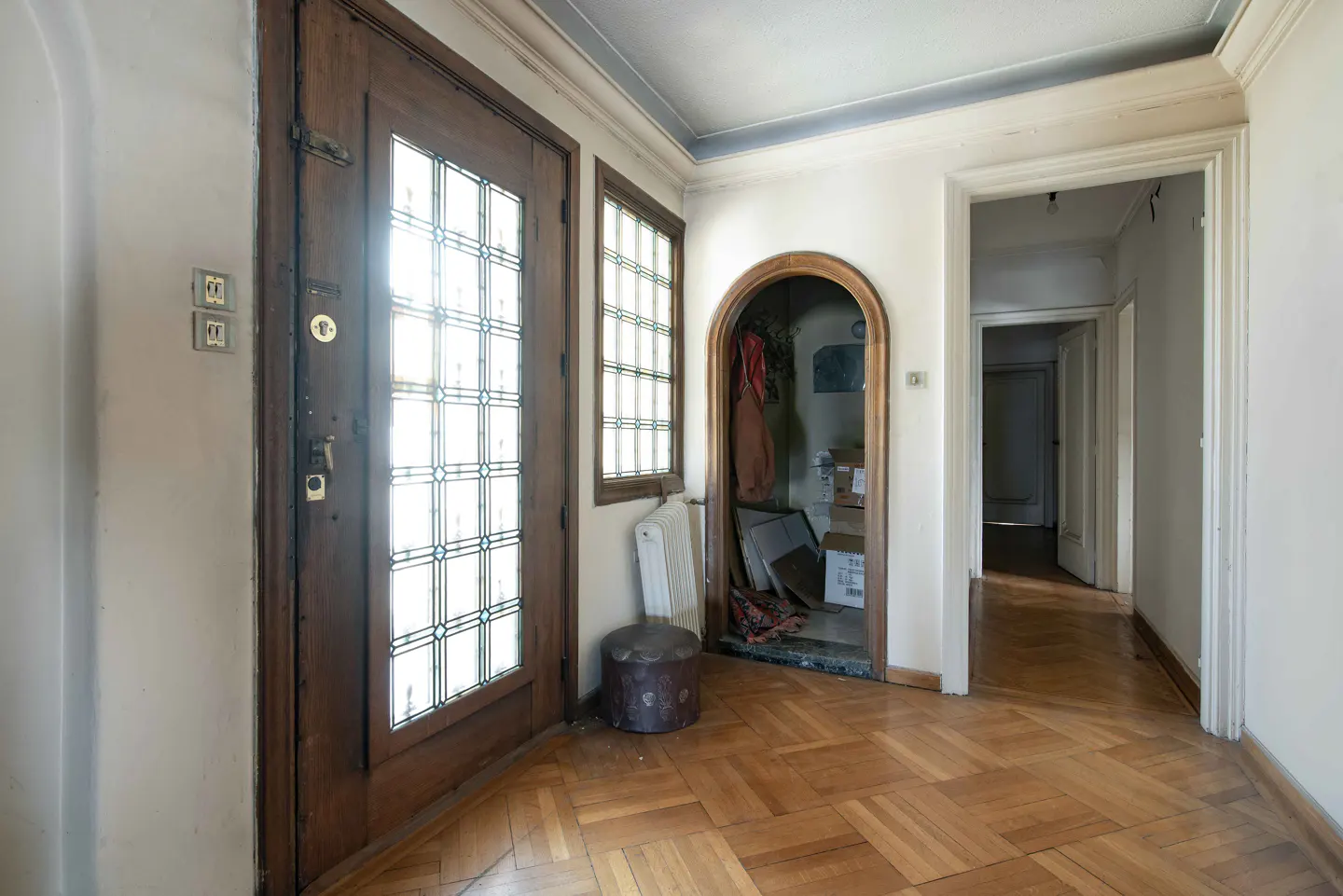 Foyer with wood parquet flooring, a glass-paneled door, and an arched doorway leading to a cluttered storage area.