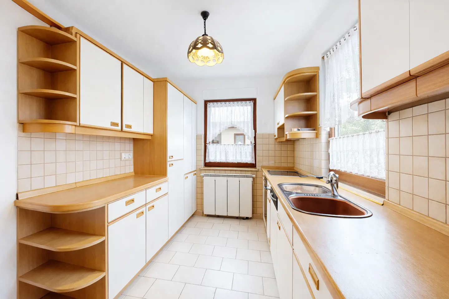 A bright kitchen with white cabinets, wood trim, and a brown sink. A window with white curtains is centered.