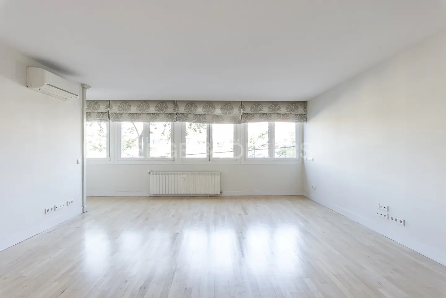 Bright, empty room with light wood floors, white walls, and large windows with patterned blinds. A radiator sits beneath the windows.