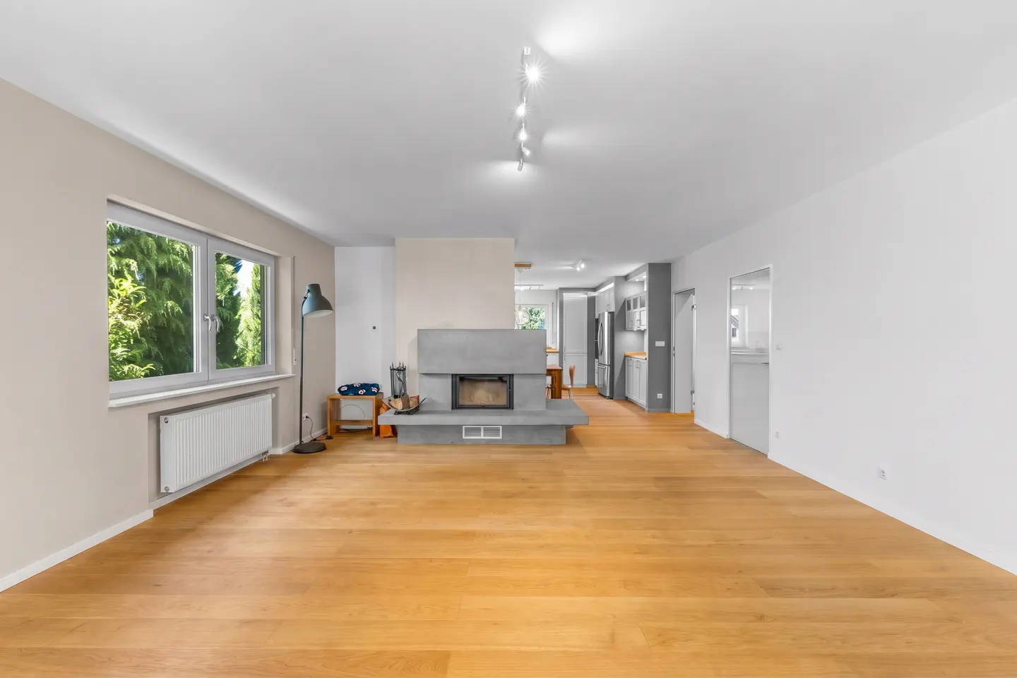 Spacious living room with light wood floors, a central grey concrete fireplace, and an open-plan kitchen in the background.