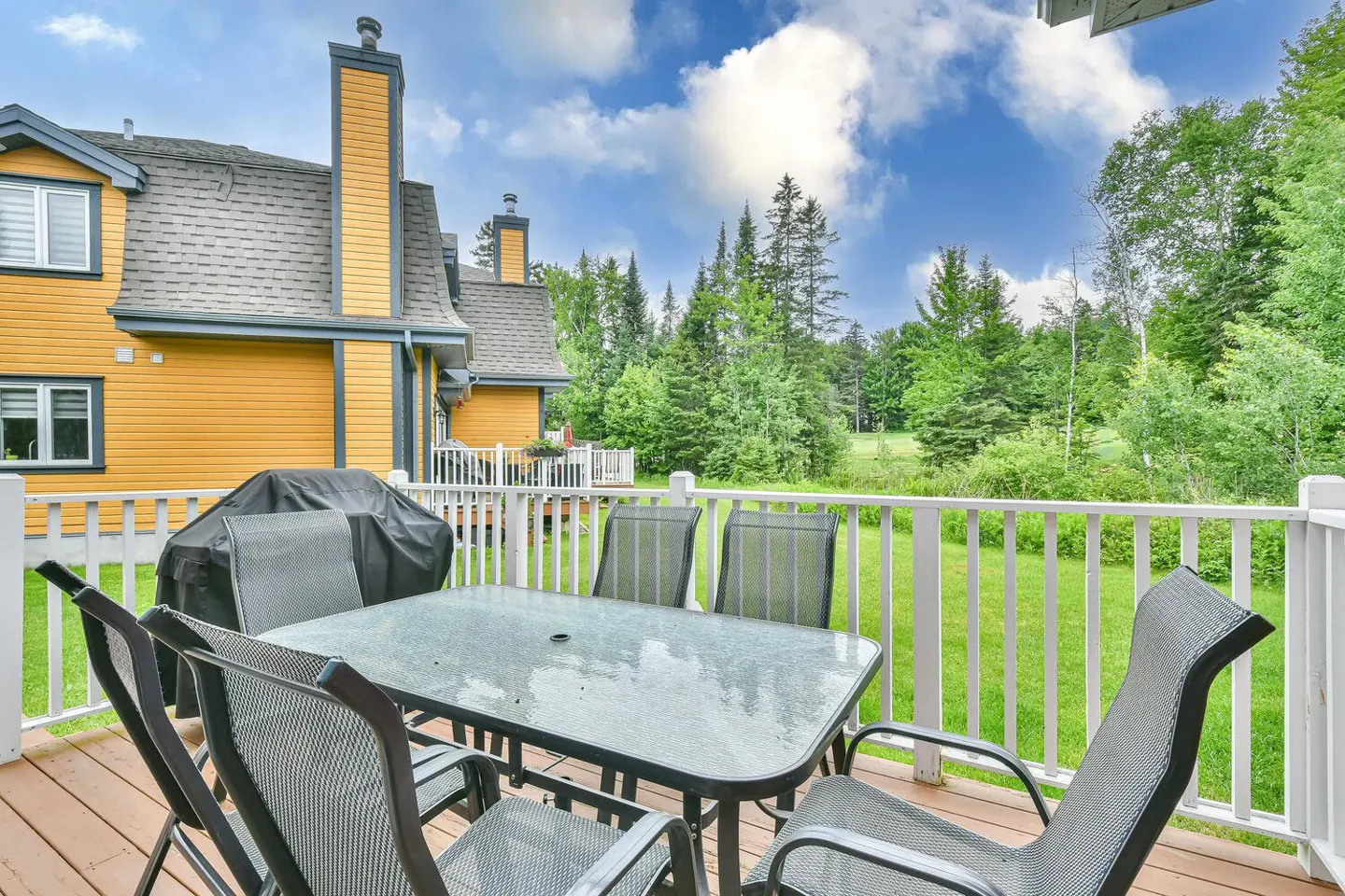 Outdoor deck with a glass table, six chairs, and a grill. A yellow house and green trees are in the background.